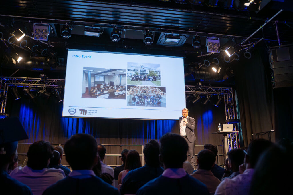 Speaker addressing the audience beneath the TU Berlin Global Production Engineering Convocation screen, photographed by an event photographer in Berlin